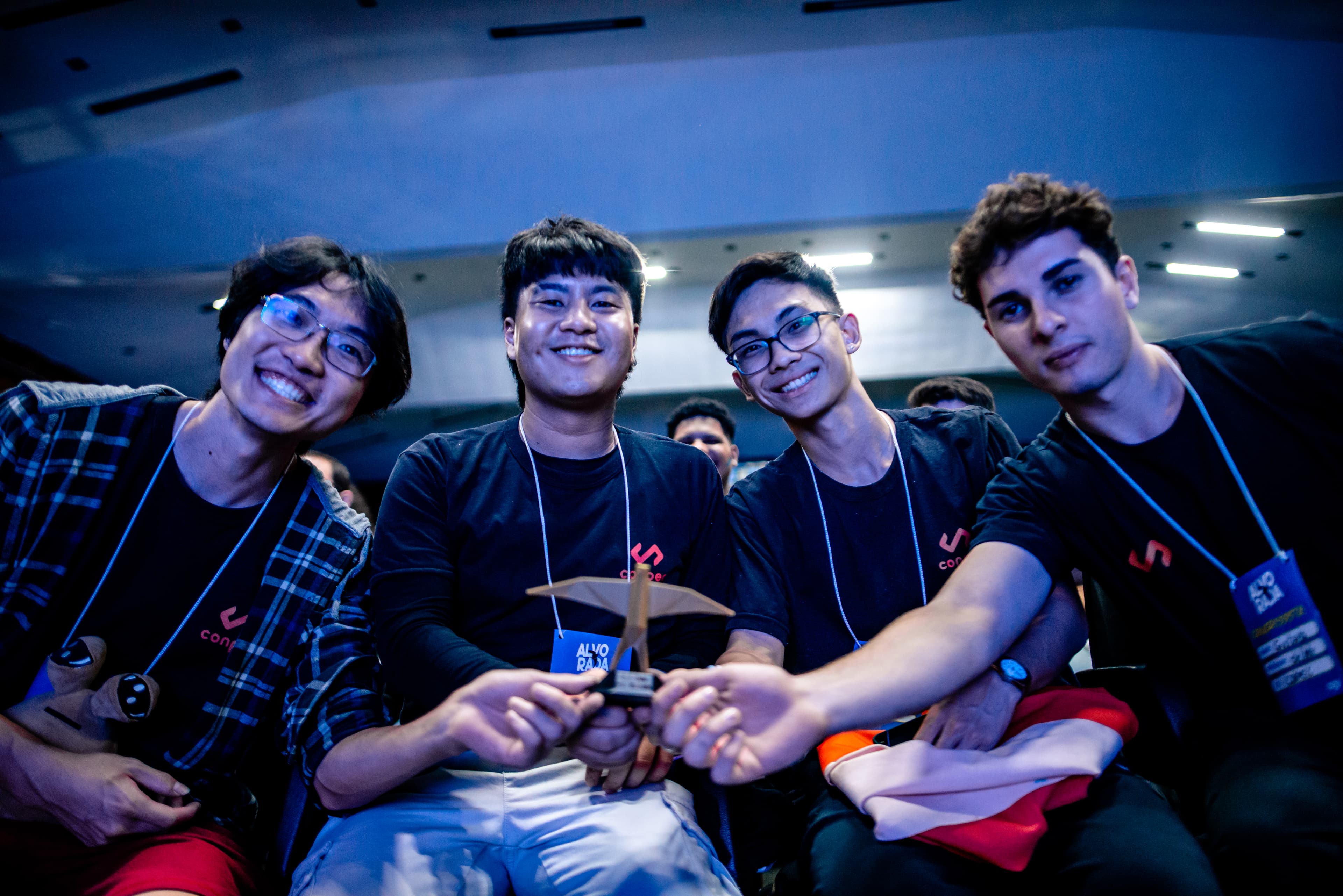 Quatro pessoas com a camisa da Conpec sorriem para a câmera, mostrando um troféu na mão. Um pou triste está no canto.