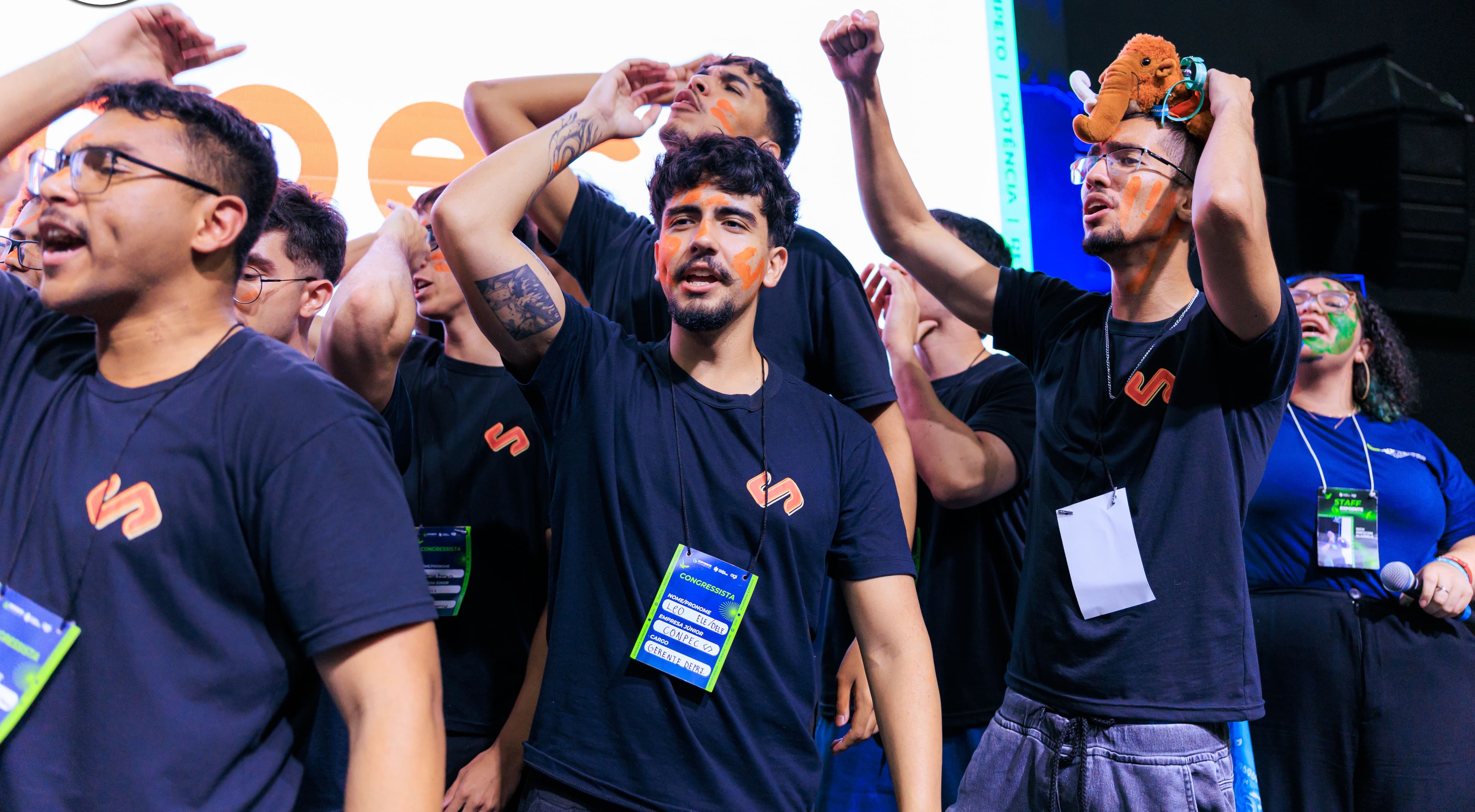 Grupo de jovens da Conpec celebrando com entusiasmo em um evento, vestindo camisetas pretas com logo laranja, crachás e pinturas faciais; um deles usa um pequeno mamute de pelúcia na cabeça.