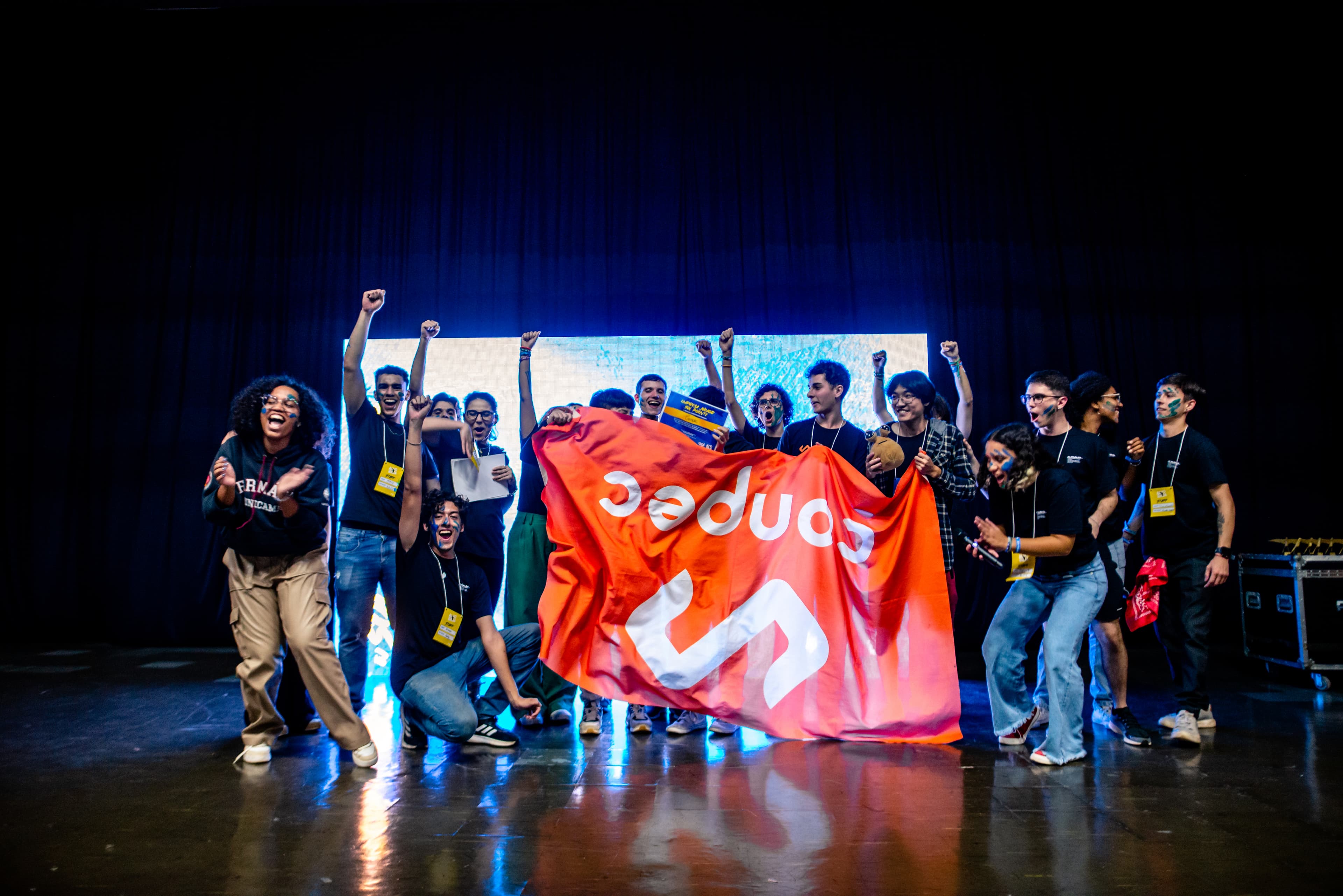 Um grupo de quinze pessoas comemora uma premiação em frente a um palco. Elas erguem a bandeira da Conpec.
