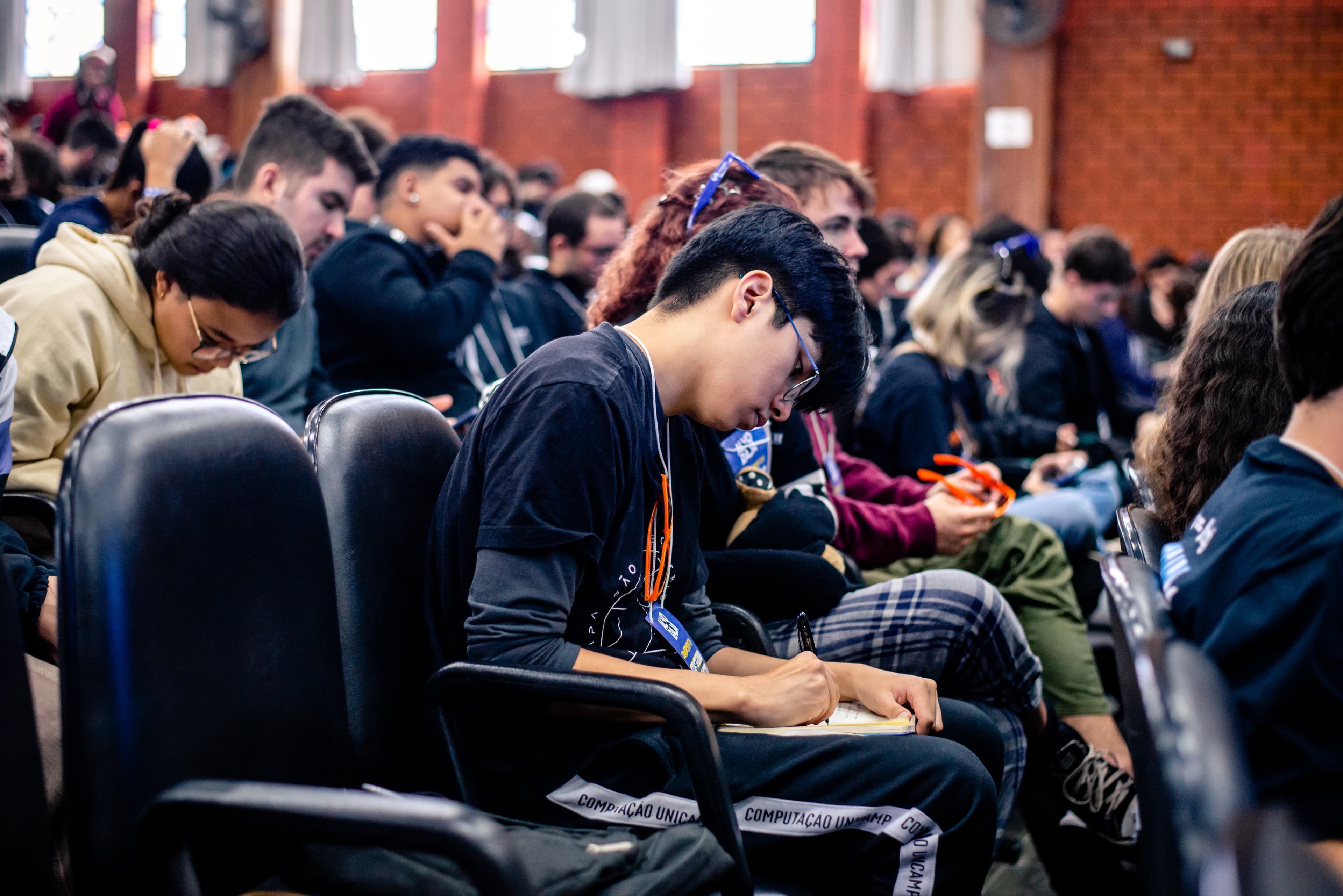 Um homem com a camisa do Movimento Empresa Júnior assiste à uma palestra, anotando num caderno.