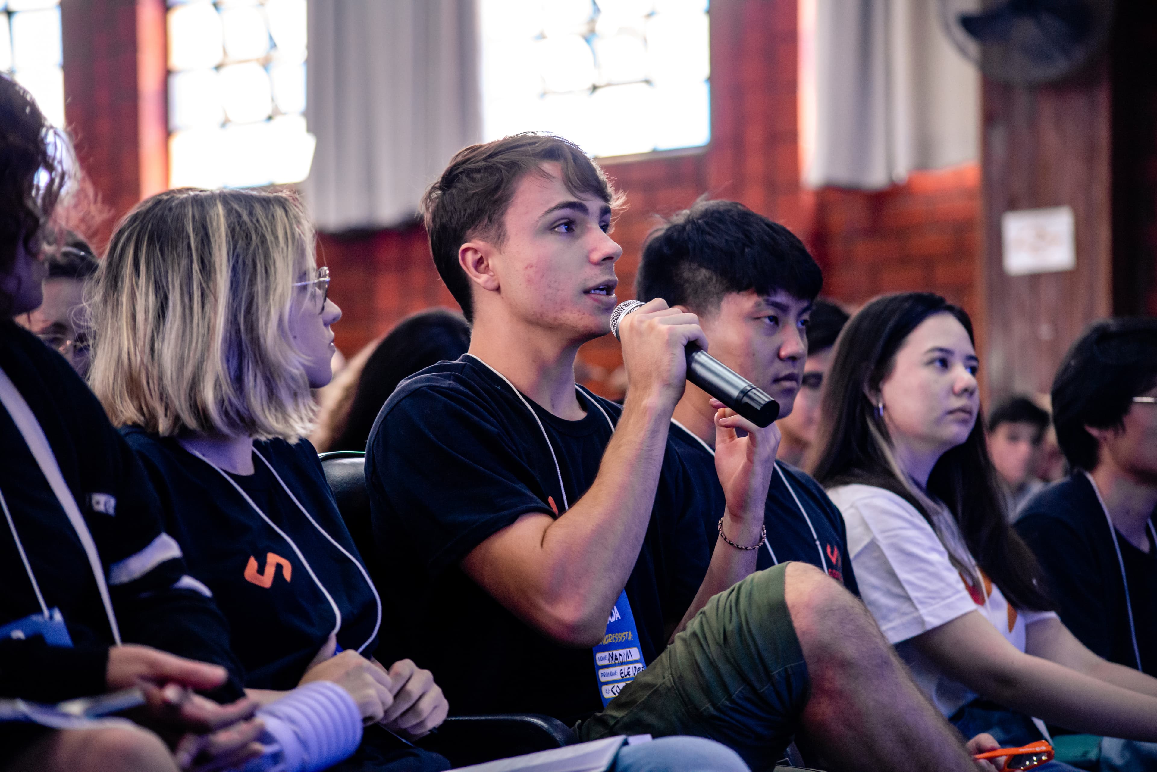 Um grupo de pessoas com a camisa da Conpec assistem uma palestra. Um homem, no meio do grupo, fala num microfone.